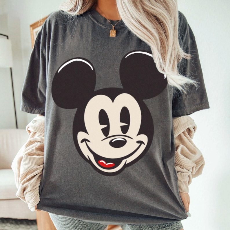 May include: Dark gray t-shirt featuring a large graphic of Mickey Mouse's face. The design includes black ears, a white face, and a smiling expression with a red mouth. The shirt has short sleeves and a relaxed fit.