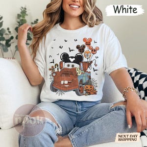 May include: White t-shirt featuring a Halloween-themed graphic. The design includes a cartoon truck with Mickey Mouse ears, balloons, pumpkins, and bats. The text "White" is printed at the top. The Cozy Nook logo is visible.