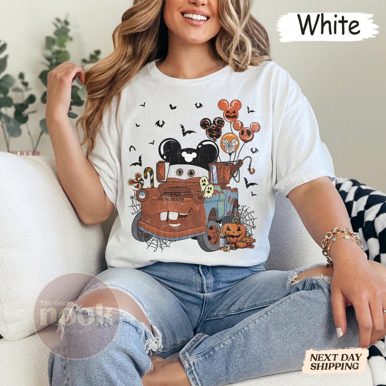 May include: White t-shirt featuring a Halloween-themed graphic. The design includes a cartoon truck with Mickey Mouse ears, balloons, pumpkins, and bats. The text "White" is printed at the top. The Cozy Nook logo is visible.