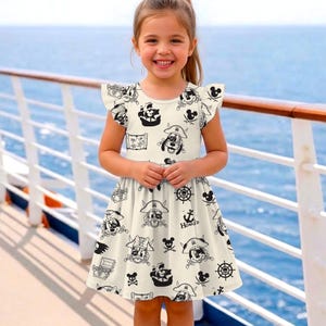 May include: A cream-colored dress with a pirate-themed print featuring cartoon characters, ships, treasure chests, and skulls. The dress has short sleeves with ruffled edges and a flared skirt. The background shows a blue ocean and a white railing.