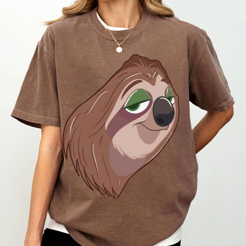 May include: Brown t-shirt featuring a cartoon sloth with brown fur, green eyes, and a black nose. The sloth has a slight smile. The shirt has short sleeves and a crew neck. The shirt is a solid brown color.