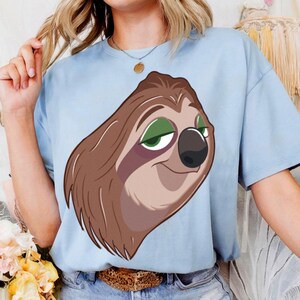 May include: Light blue t-shirt featuring a cartoon sloth with brown fur, green eyes, and a black nose. The sloth is smiling. The shirt is made of a soft material.
