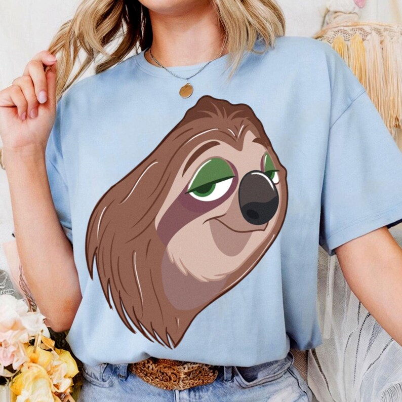 May include: Light blue t-shirt featuring a cartoon sloth with brown fur, green eyes, and a black nose. The sloth is smiling. The shirt is made of a soft material.