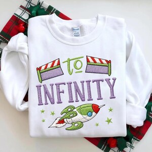 Puede incluir: Sudadera blanca de cuello redondo con dise&ntilde;o bordado. El dise&ntilde;o incluye la frase "TO INFINITY" en morado, un cohete verde y rojo, y estrellas verdes. La sudadera est&aacute; sobre una tela a cuadros roja y verde.
