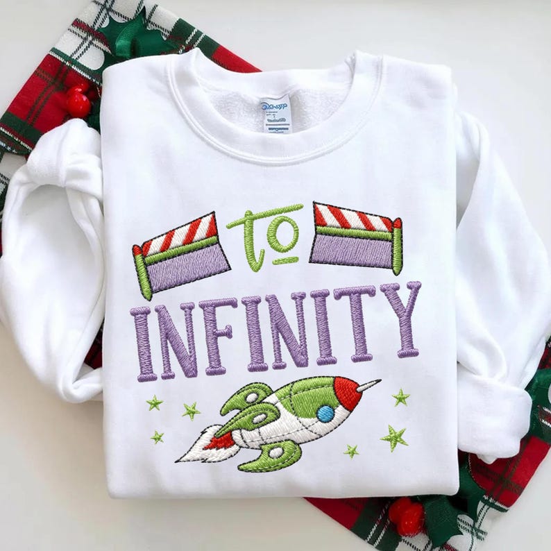 Puede incluir: Sudadera blanca de cuello redondo con dise&ntilde;o bordado. El dise&ntilde;o incluye la frase "TO INFINITY" en morado, un cohete verde y rojo, y estrellas verdes. La sudadera est&aacute; sobre una tela a cuadros roja y verde.