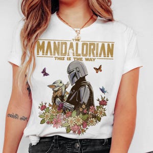 May include: White t-shirt featuring a graphic of The Mandalorian and Grogu surrounded by flowers and butterflies. The text "THE MANDALORIAN" and "THIS IS THE WAY" are printed across the top.