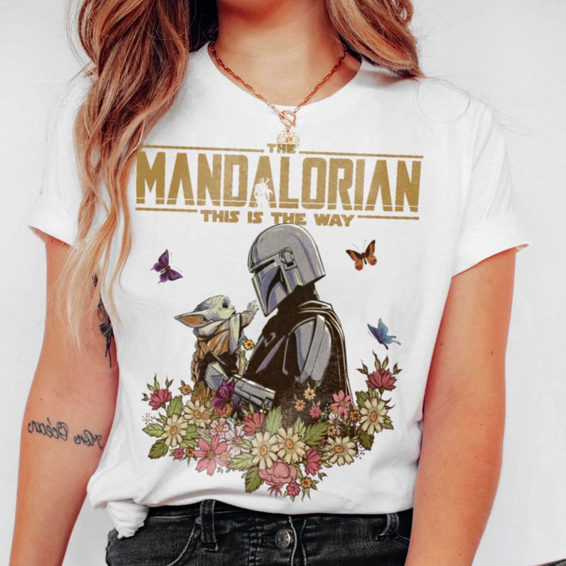 May include: White t-shirt featuring a graphic of The Mandalorian and Grogu surrounded by flowers and butterflies. The text "THE MANDALORIAN" and "THIS IS THE WAY" are printed across the top.