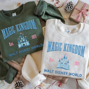 May include: Two crewneck sweatshirts, one olive green and one cream, embroidered with "MAGIC KINGDOM" and "WALT DISNEY WORLD." The design includes a castle graphic and the year "1971." Festive holiday decorations surround the sweatshirts.