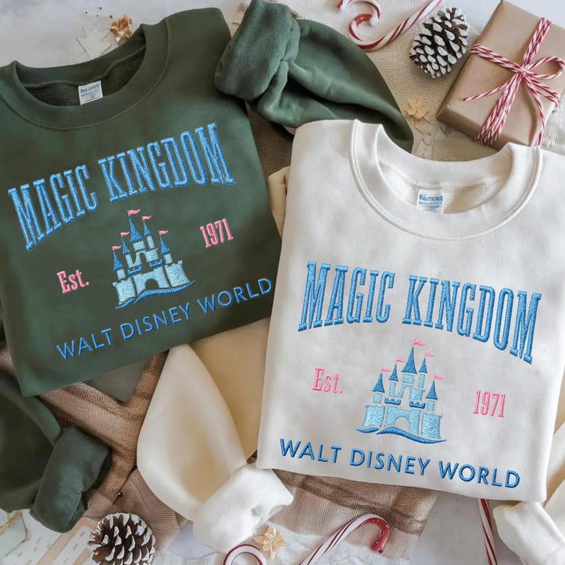 May include: Two crewneck sweatshirts, one olive green and one cream, embroidered with "MAGIC KINGDOM" and "WALT DISNEY WORLD." The design includes a castle graphic and the year "1971." Festive holiday decorations surround the sweatshirts.