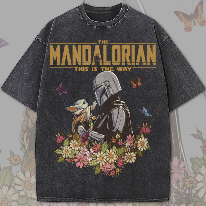 May include: Dark gray t-shirt featuring a graphic of The Mandalorian and Grogu surrounded by flowers and butterflies. The text "THE MANDALORIAN" and "THIS IS THE WAY" are printed across the top in gold.