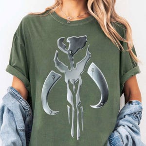 May include: An olive green t-shirt displaying a metallic silver emblem. The emblem is a symmetrical design with curved and angular elements. The shirt has short sleeves and a relaxed fit, suitable for casual wear.