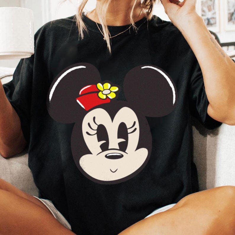 Peut inclure: T-shirt noir avec un grand motif de Minnie Mouse. Le design pr&eacute;sente son visage avec des oreilles noires, un n&oelig;ud rouge avec une fleur jaune et une expression souriante. Le t-shirt est de style d&eacute;contract&eacute;.