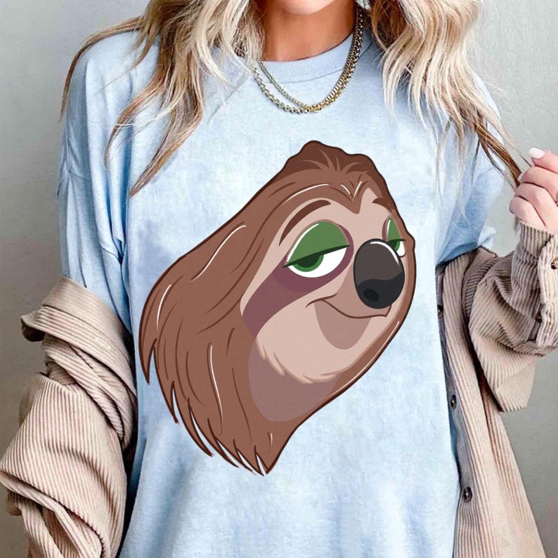 May include: Light blue t-shirt featuring a cartoon sloth with brown fur, a black nose, and green eyes. The sloth is smiling. The shirt is made of a soft material and is suitable for casual wear.
