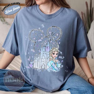 May include: Blue jean-colored t-shirt featuring a graphic of Elsa from Frozen, a castle, and a Mickey Mouse-shaped design with stars and hearts. The shirt has the words "Comfort Colors" and "Blue Jean" in a circular design.