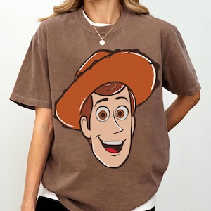 May include: Brown t-shirt featuring a cartoon graphic of a smiling cowboy character with a brown hat. The graphic has a white outline. The shirt has short sleeves and a crew neck. The shirt is a solid brown color.