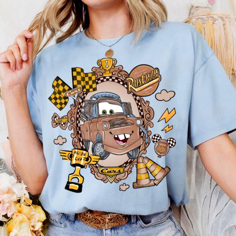 May include: Light blue t-shirt featuring a cartoon image of Tow Mater from the movie Cars. The graphic includes a brown frame, checkered flags, and the text "Rusteze" and "Cars". Additional elements include a trophy, lightning bolts, and traffic cones.