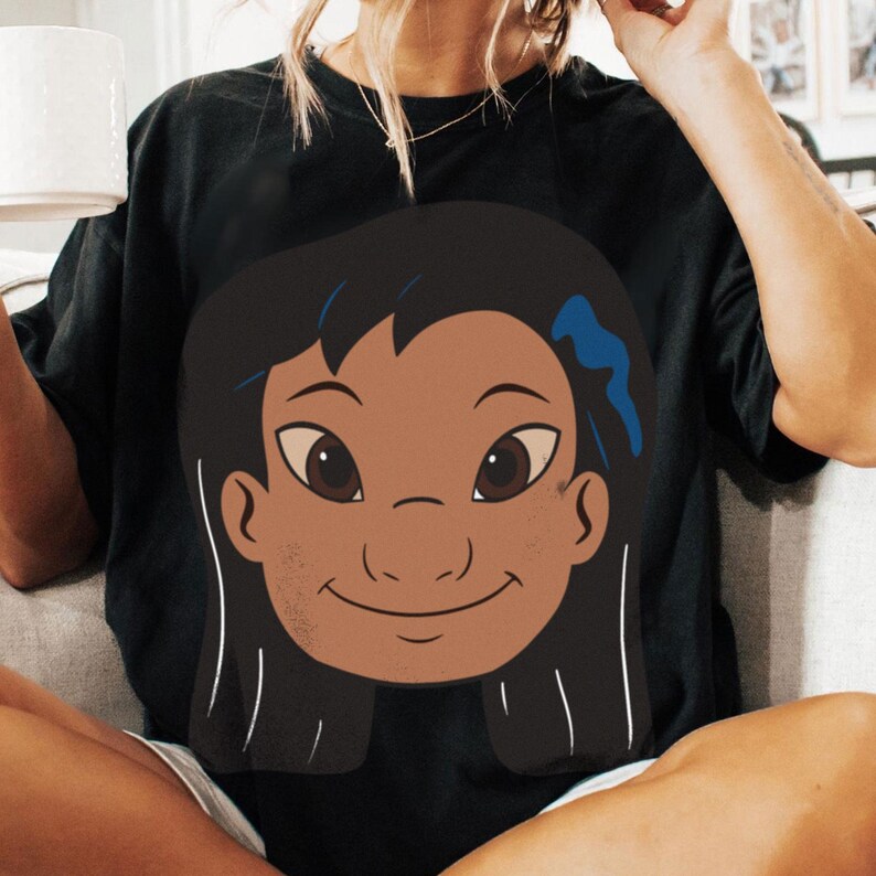 Puede incluir: Camiseta negra con una ilustraci&oacute;n de dibujos animados de una ni&ntilde;a sonriente con cabello oscuro, ojos marrones y un accesorio para el cabello azul. El gr&aacute;fico est&aacute; centrado en la parte delantera de la camiseta. Ropa casual.