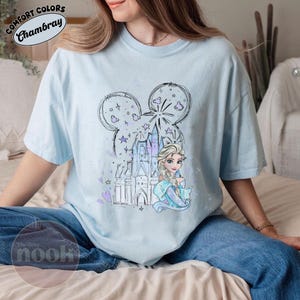 May include: Light blue t-shirt featuring a cartoon illustration of a princess and a castle, with a Mickey Mouse-shaped design above. The shirt has the text "Comfort Colors Chambray" and "The Cozy Nook".