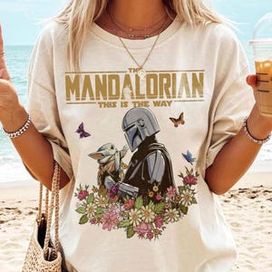 May include: Cream-colored t-shirt featuring the text "THE MANDALORIAN" above an illustration of the Mandalorian and Grogu surrounded by flowers and butterflies. The phrase "THIS IS THE WAY" is printed below the title. The design is centered on the shirt.