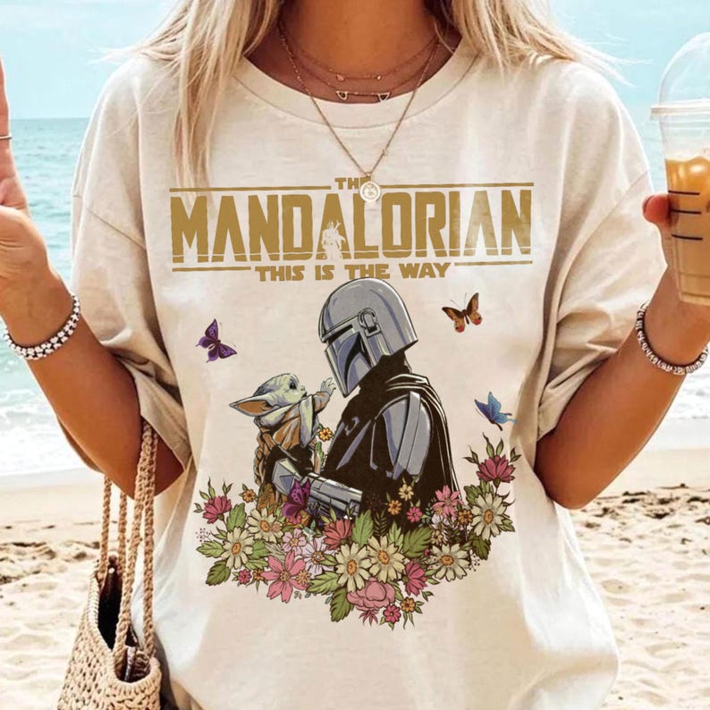 May include: Cream-colored t-shirt featuring the text "THE MANDALORIAN" above an illustration of the Mandalorian and Grogu surrounded by flowers and butterflies. The phrase "THIS IS THE WAY" is printed below the title. The design is centered on the shirt.