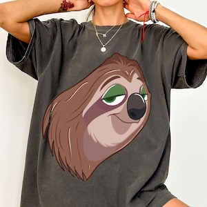 May include: Dark gray t-shirt featuring a cartoon sloth face with brown fur, green eyes, and a black nose. The sloth has a relaxed expression. The shirt has short sleeves and a slightly faded appearance, suggesting a vintage style.