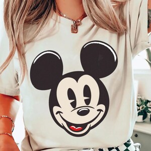 May include: Cream-colored t-shirt featuring a large graphic of Mickey Mouse's face. The design includes black ears, a black outline, a white face, and a red mouth. The shirt is a casual style.