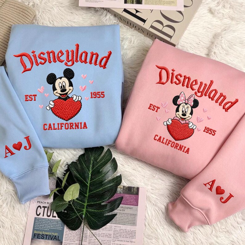 Puede incluir: Dos sudaderas, una celeste y otra rosa, bordadas con "Disneyland" en escritura roja. Cada una presenta a Mickey o Minnie Mouse sosteniendo un coraz&oacute;n rojo, con "EST 1955 CALIFORNIA" debajo. Las mangas tienen iniciales. Una revista y vegetaci&oacute;n en primer plano.
