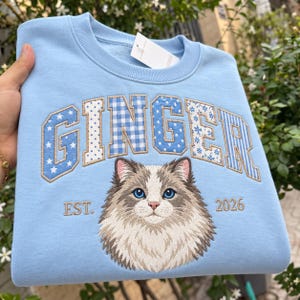 May include: A light blue crewneck sweatshirt featuring the word GINGER in large, multi-patterned blue and white embroidered letters. Below the text is an embroidered portrait of a fluffy cat with blue eyes, flanked by the text EST. and 2026.