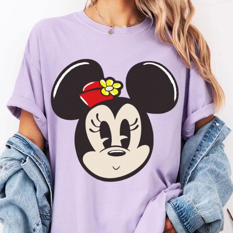Peut inclure: T-shirt lilas avec un grand motif de Minnie Mouse. Le visage du personnage est en noir et blanc, avec un n&oelig;ud rouge et une fleur jaune. Les manches du t-shirt sont retrouss&eacute;es.