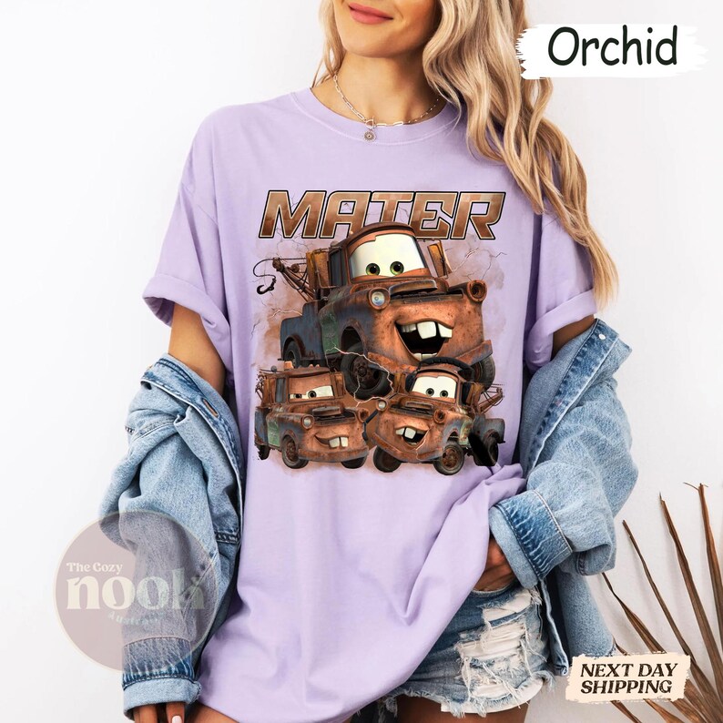 May include: Lilac t-shirt with a graphic of the character Mater from the film Cars. The word "MATER" is printed above the graphic in a brown, distressed font. The shirt has a relaxed fit and rolled sleeves.