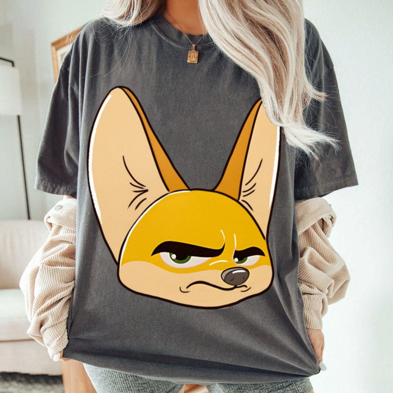 May include: Dark gray t-shirt featuring a large, cartoonish fennec fox face. The fox has large, pointed ears, a yellow and orange face, and a slightly grumpy expression with green eyes. The shirt is a casual, oversized style.
