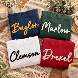May include: Four crewneck sweatshirts featuring embroidered script lettering. The navy sweatshirt has yellow text reading Baylor, the green has white text reading Marlow, the white has black text reading Clemson, and the red has white text reading Drexel.