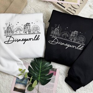 May include: Two sweatshirts, one white and one black, featuring a line drawing of a castle, tree, building, and globe. The word "Disneyworld" is written in a cursive font below the graphic. The sweatshirts are folded on a white surface.