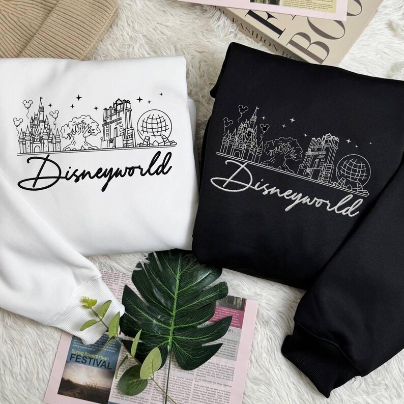 May include: Two sweatshirts, one white and one black, featuring a line drawing of a castle, tree, building, and globe. The word "Disneyworld" is written in a cursive font below the graphic. The sweatshirts are folded on a white surface.