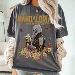 May include: Dark gray t-shirt featuring a graphic of The Mandalorian and Grogu surrounded by flowers and butterflies. The text "THE MANDALORIAN THIS IS THE WAY" is printed across the top in gold lettering. A unique and stylish tee.