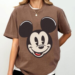 May include: Brown t-shirt featuring a large graphic of Mickey Mouse's face. The cartoon character's face is in black, beige, and red. The shirt has short sleeves and a crew neck, with a white undershirt visible.