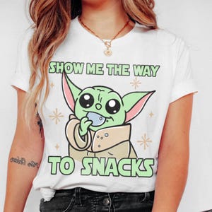 Peut inclure: T-shirt blanc avec un personnage de dessin anim&eacute; vert tenant une collation. Le texte "SHOW ME THE WAY TO SNACKS" est imprim&eacute; au-dessus et en dessous du personnage en vert. Le design comprend des &eacute;toiles.