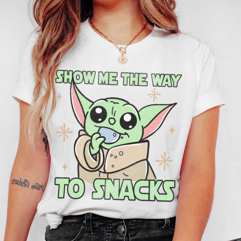 Peut inclure: T-shirt blanc avec un personnage de dessin anim&eacute; vert tenant une collation. Le texte "SHOW ME THE WAY TO SNACKS" est imprim&eacute; au-dessus et en dessous du personnage en vert. Le design comprend des &eacute;toiles.