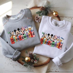May include: Two sweatshirts, one gray and one white, each featuring a colorful Disney World graphic. The graphic includes Mickey Mouse, Minnie Mouse, Donald Duck, and other characters. The word "Disneyworld" is printed in a rainbow-colored font.