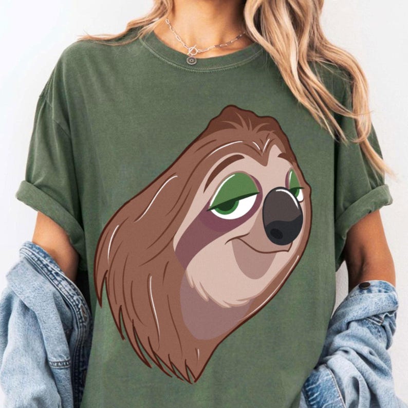 May include: Olive green t-shirt featuring a cartoon sloth with brown fur, green eyes, and a black nose. The sloth has a relaxed expression. The shirt has short sleeves and a casual fit.