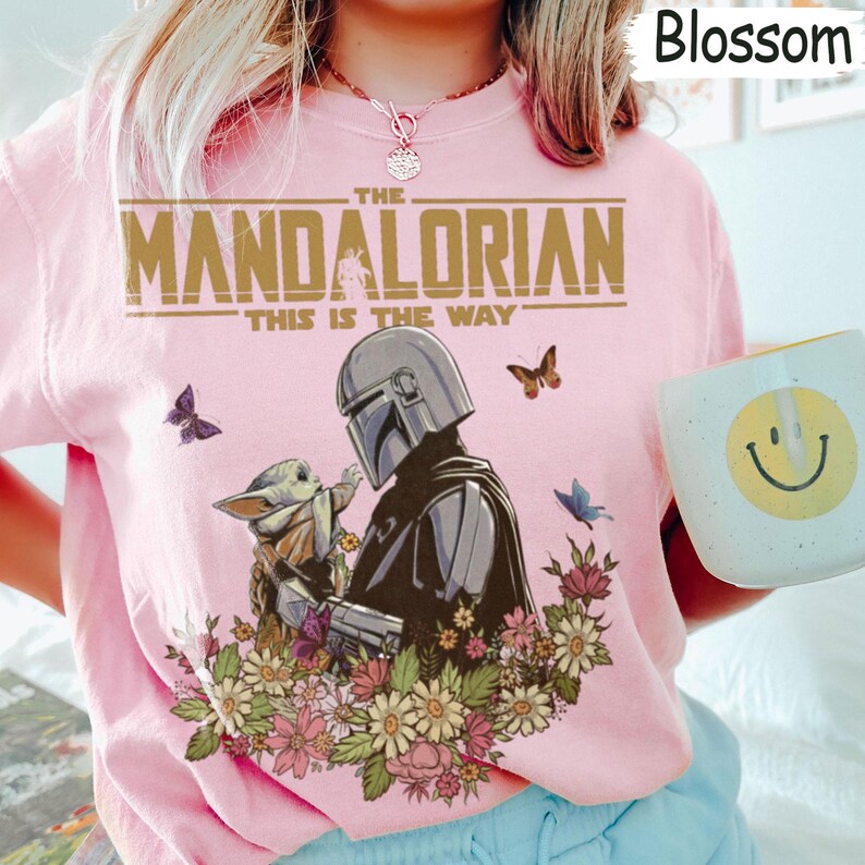 May include: A light pink t-shirt featuring a graphic of The Mandalorian and Grogu, surrounded by flowers and butterflies. The text "THE MANDALORIAN THIS IS THE WAY" is printed across the top. A white mug with a yellow smiley face is visible.
