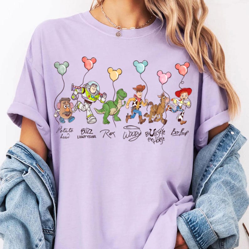 May include: A lilac t-shirt with a cartoon graphic of Toy Story characters holding balloons. The design includes Potato Head, Buzz Lightyear, Rex, Woody, Bullseye the Horse, and Bo Peep. The characters' names are written below them.