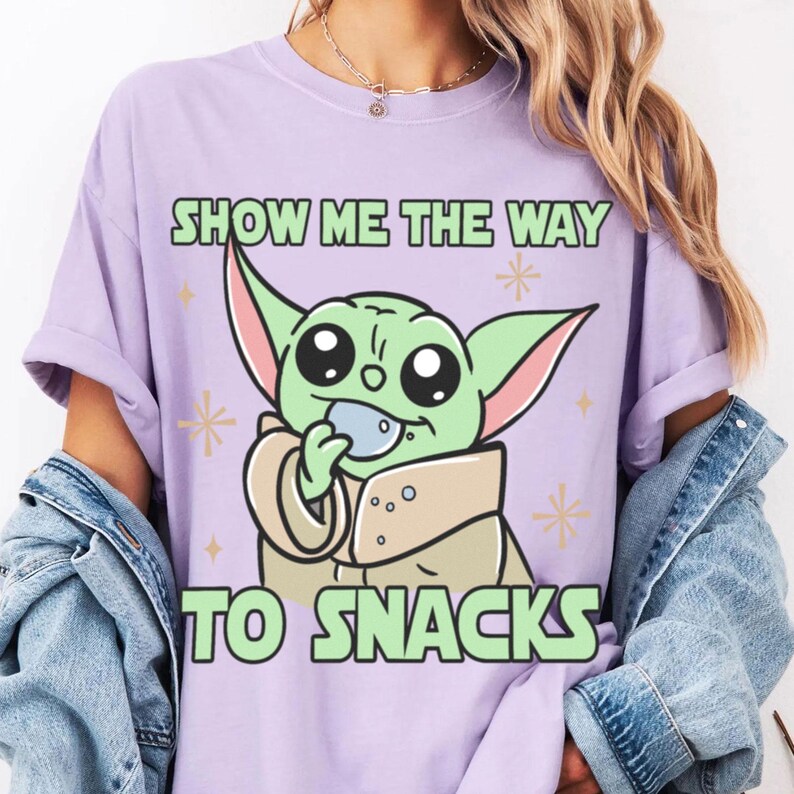 Peut inclure: T-shirt lilas avec un personnage de dessin anim&eacute; vert tenant une collation. Le texte "SHOW ME THE WAY TO SNACKS" est imprim&eacute; au-dessus et en dessous du personnage en lettres vertes. Le design comprend des &eacute;toiles.