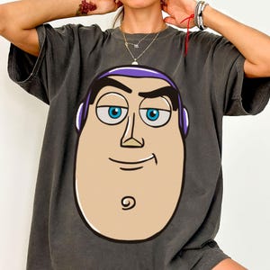 May include: Dark gray t-shirt featuring a large cartoon image of Buzz Lightyear's face. The graphic includes his blue eyes, purple headpiece, and a smiling expression. The shirt has short sleeves and a relaxed fit.