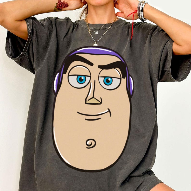 May include: Dark gray t-shirt featuring a large cartoon image of Buzz Lightyear's face. The graphic includes his blue eyes, purple headpiece, and a smiling expression. The shirt has short sleeves and a relaxed fit.