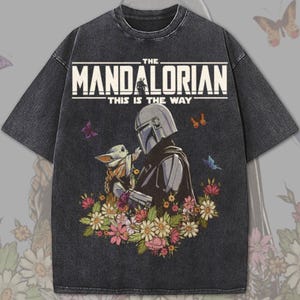May include: Dark gray t-shirt featuring a graphic of The Mandalorian and Grogu surrounded by flowers. The text "THE MANDALORIAN" and "THIS IS THE WAY" are printed across the top in white. Butterflies are scattered around the design.