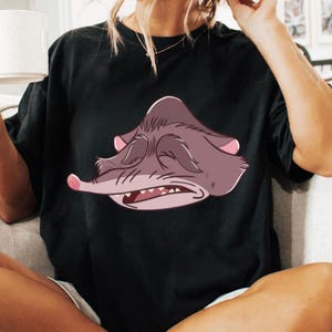Peut inclure: T-shirt noir avec une illustration de dessin anim&eacute; d'une cr&eacute;ature grincheuse, violet-gris avec des accents roses. La cr&eacute;ature a les yeux ferm&eacute;s, un long nez et des dents visibles. Le t-shirt est de style d&eacute;contract&eacute;.