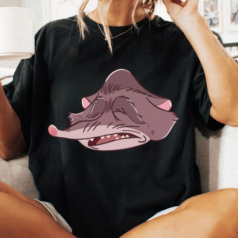 Peut inclure: T-shirt noir avec une illustration de dessin anim&eacute; d'une cr&eacute;ature grincheuse, violet-gris avec des accents roses. La cr&eacute;ature a les yeux ferm&eacute;s, un long nez et des dents visibles. Le t-shirt est de style d&eacute;contract&eacute;.