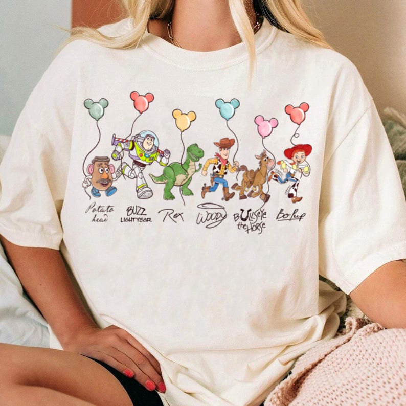 May include: A cream-coloured t-shirt with a vibrant graphic of Toy Story characters, each holding Mickey Mouse-shaped balloons. The design includes Mr. Potato Head, Buzz Lightyear, Rex, Woody, Bullseye the Horse, and Bo Peep, with their names underneath.