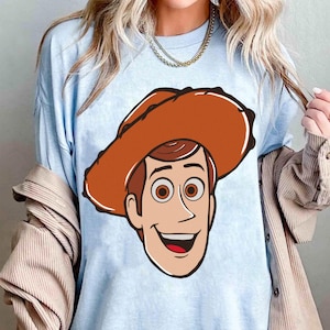 May include: Light blue t-shirt featuring a cartoon illustration of a smiling cowboy character with a brown hat and hair, and large brown eyes. The graphic is centered on the shirt.